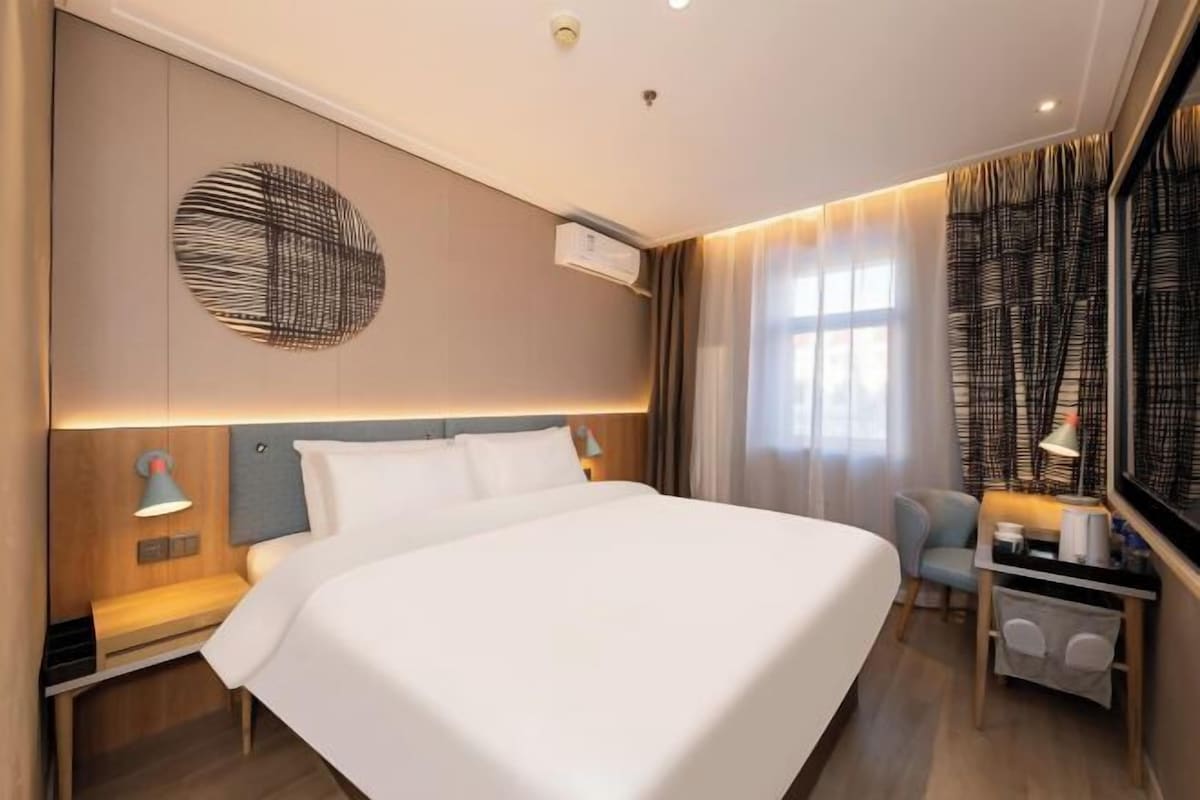 A hotel room with a large bed, a desk with a lamp, a wall-mounted air conditioner, and a window with curtains.