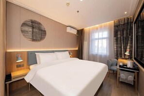 Quarto de hotel com uma cama grande, uma mesa com luminária, ar-condicionado fixado na parede e uma janela com cortinas.