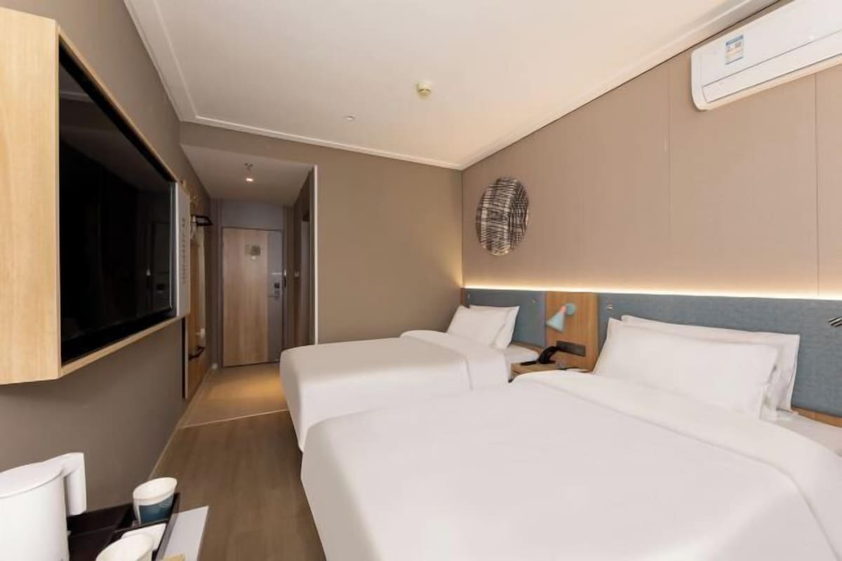 A hotel room with two beds, a TV, a wall-mounted artwork, and a bedside table with a lamp.