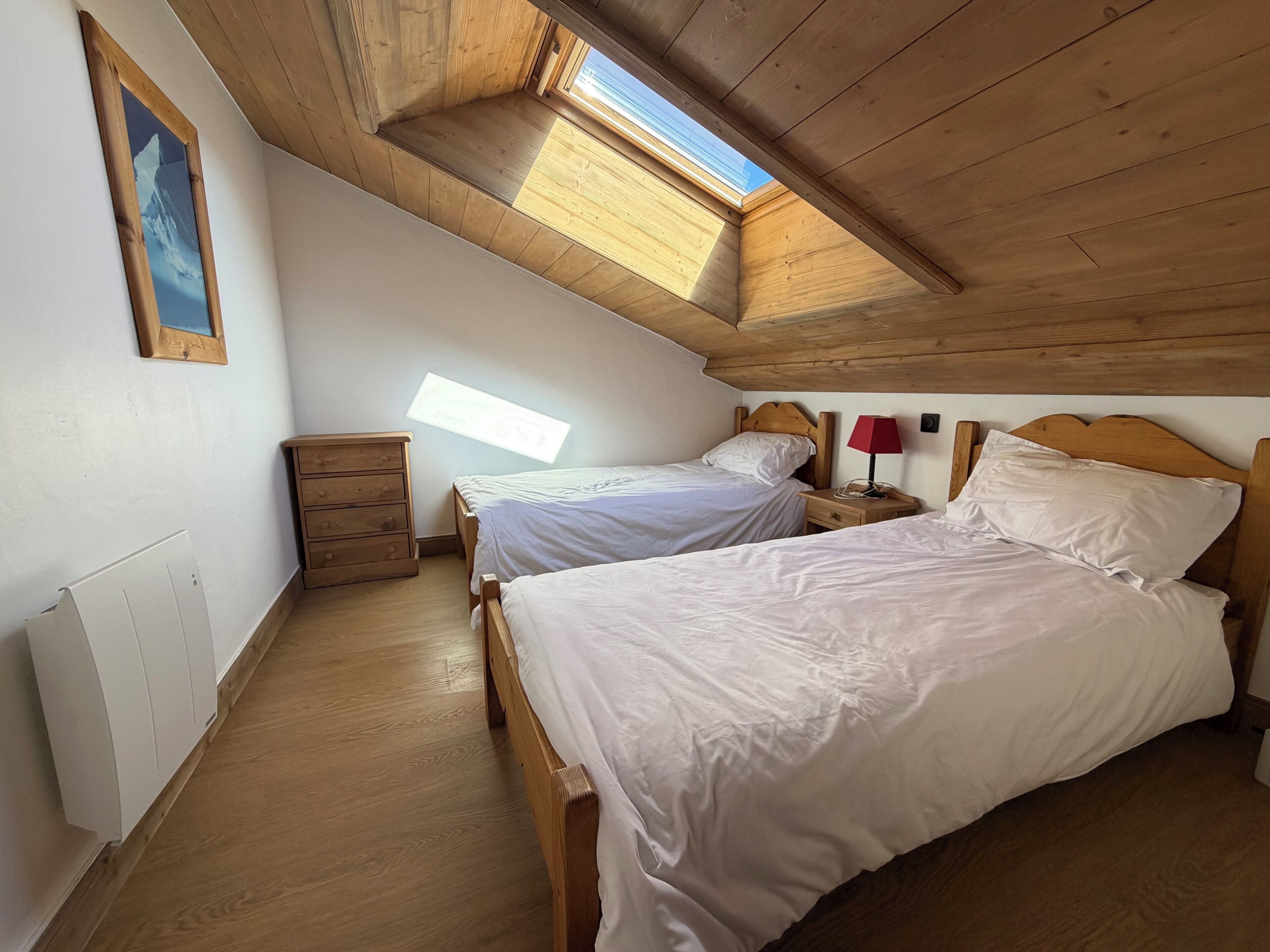 A bedroom with two beds, wooden flooring, a dresser, a radiator, and a skylight.