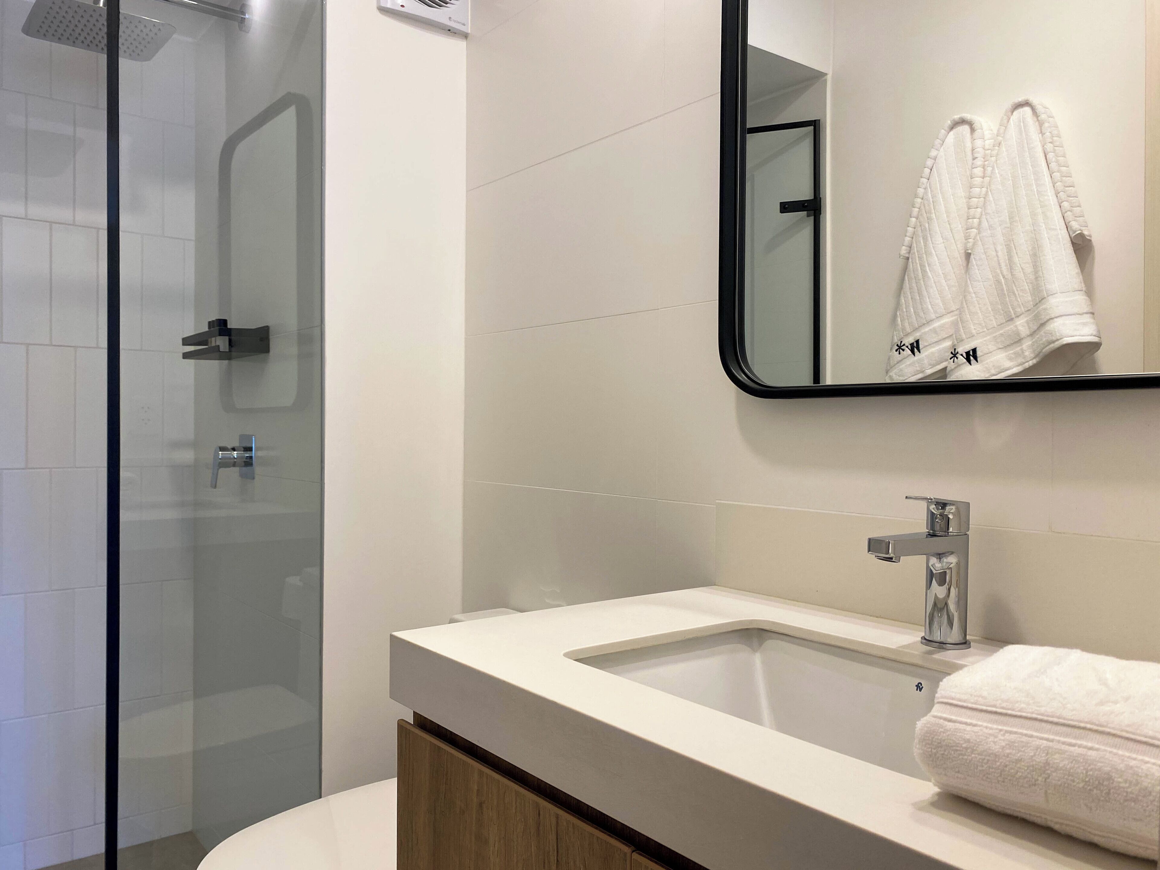 Une salle de bain moderne avec une douche à l’italienne en verre, un lavabo blanc avec un miroir au-dessus, et une serviette accrochée au mur.