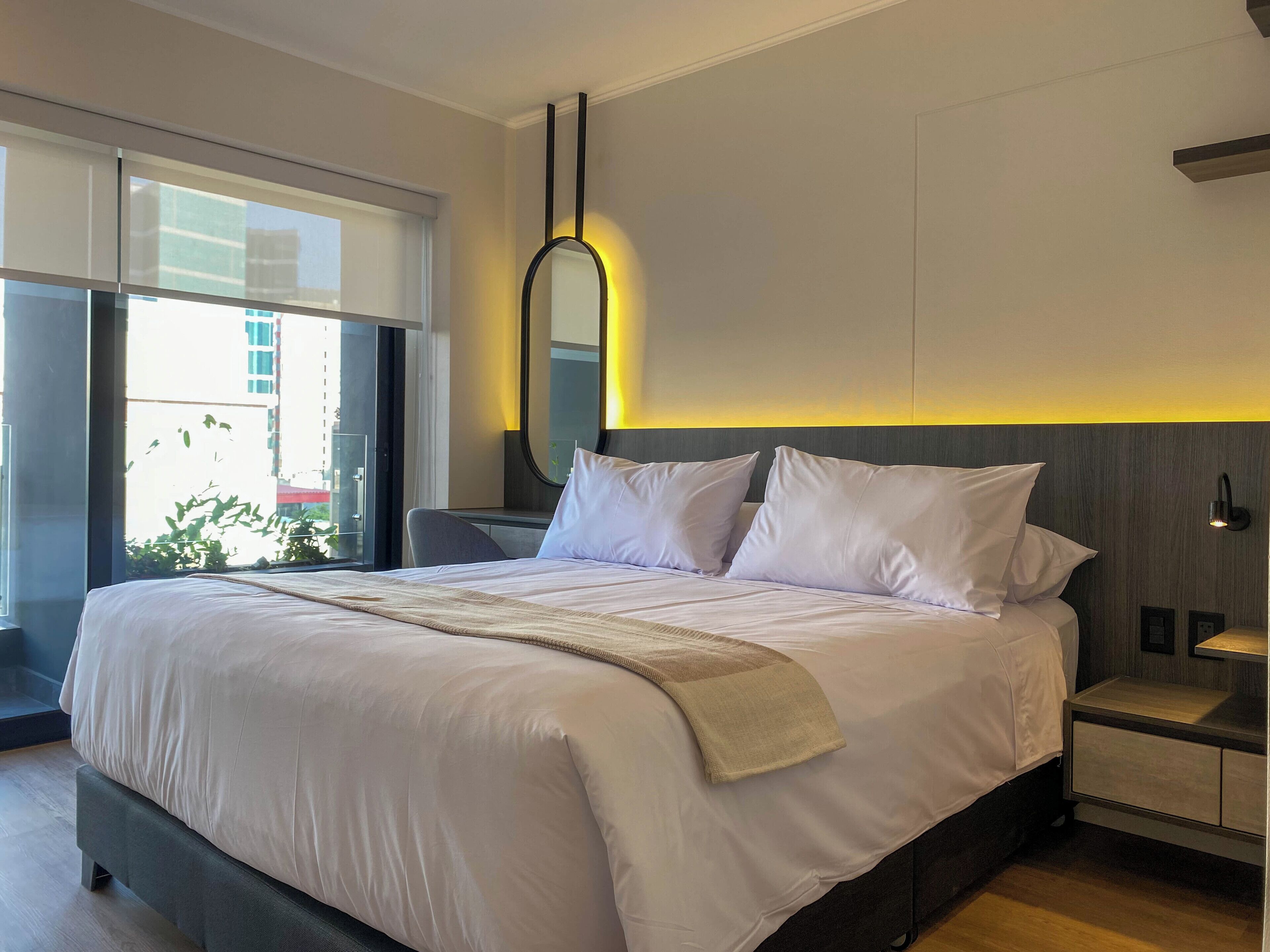 A modern hotel room with a large bed, a bedside table, a mirror, and a view of the outdoors.