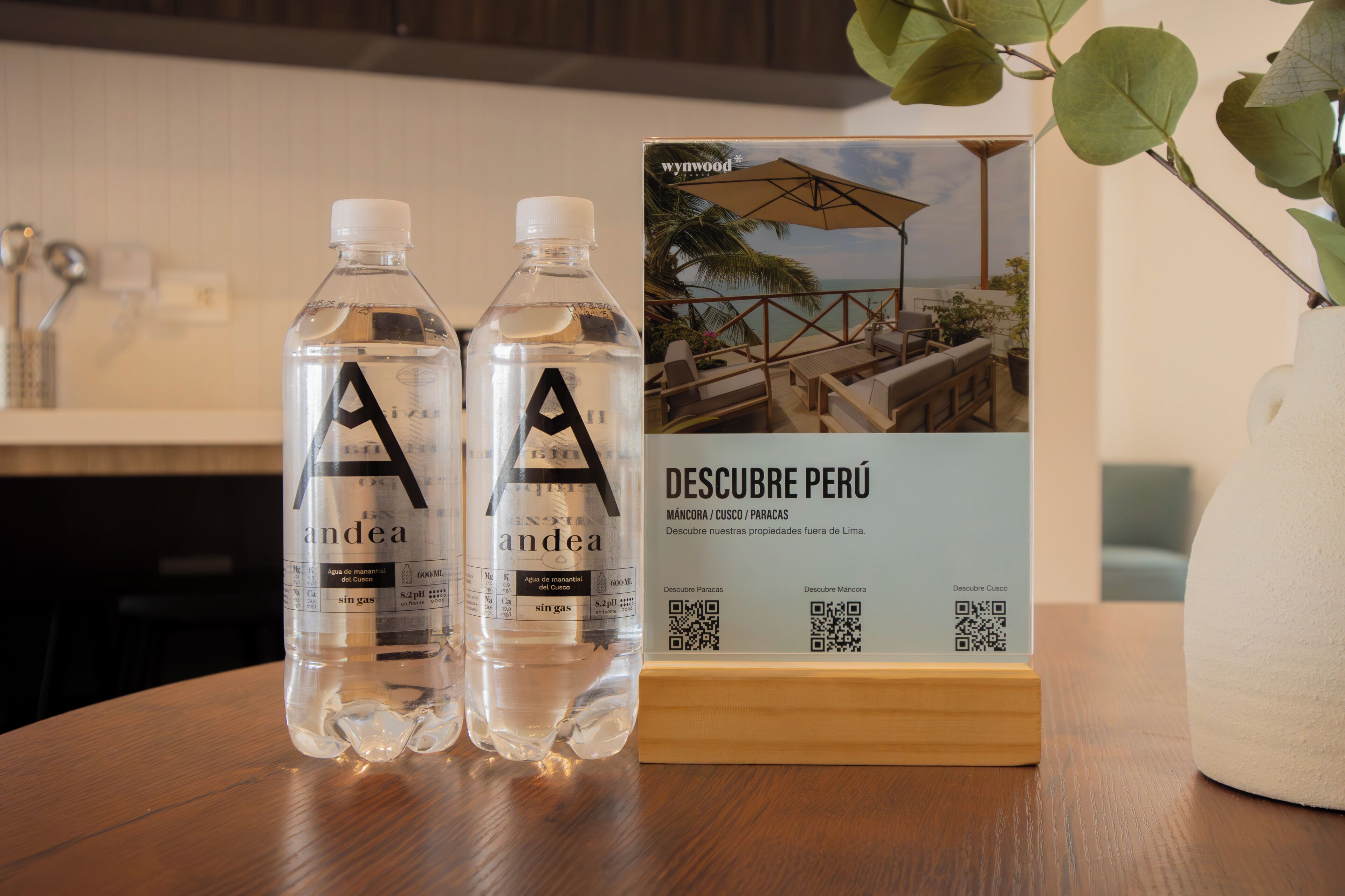 Twee flessen water met de aanduiding 'andea' op een houten tafel, een brochure getiteld 'DESCUBRE PERÚ' met QR-codes, en een potplant op de achtergrond.