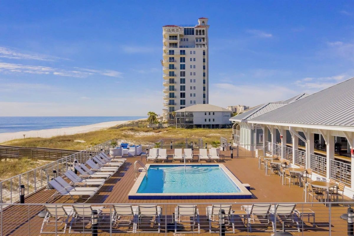 Ein Poolbereich mit Liegestühlen und Blick auf den Strand sowie ein hohes Gebäude im Hintergrund.