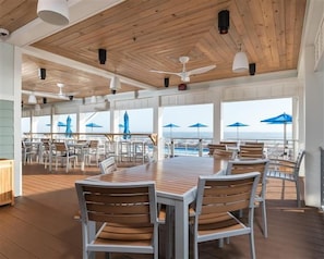 Un vaste espace extérieur aménagé pour les repas, avec des tables et des chaises en bois, de grandes fenêtres et des parasols bleus.