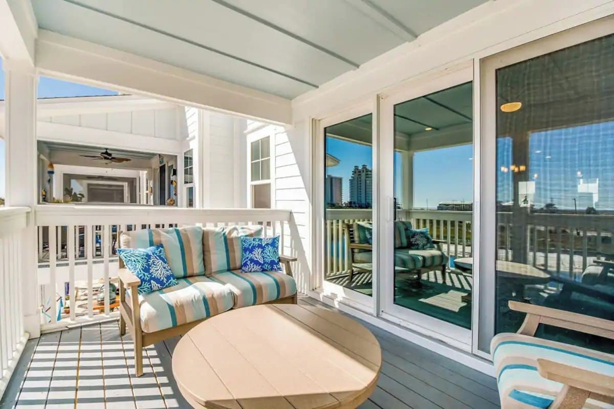A covered outdoor deck with a sofa and a round table, a glass door leading to a balcony, and a view of the cityscape.