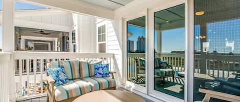 A covered outdoor deck with a sofa and a round table, a glass door leading to a balcony, and a view of the cityscape.
