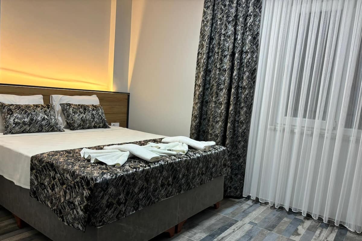 A hotel room with a bed, pillows, a bedside table, a lamp, a window with curtains, and a view of the outside.