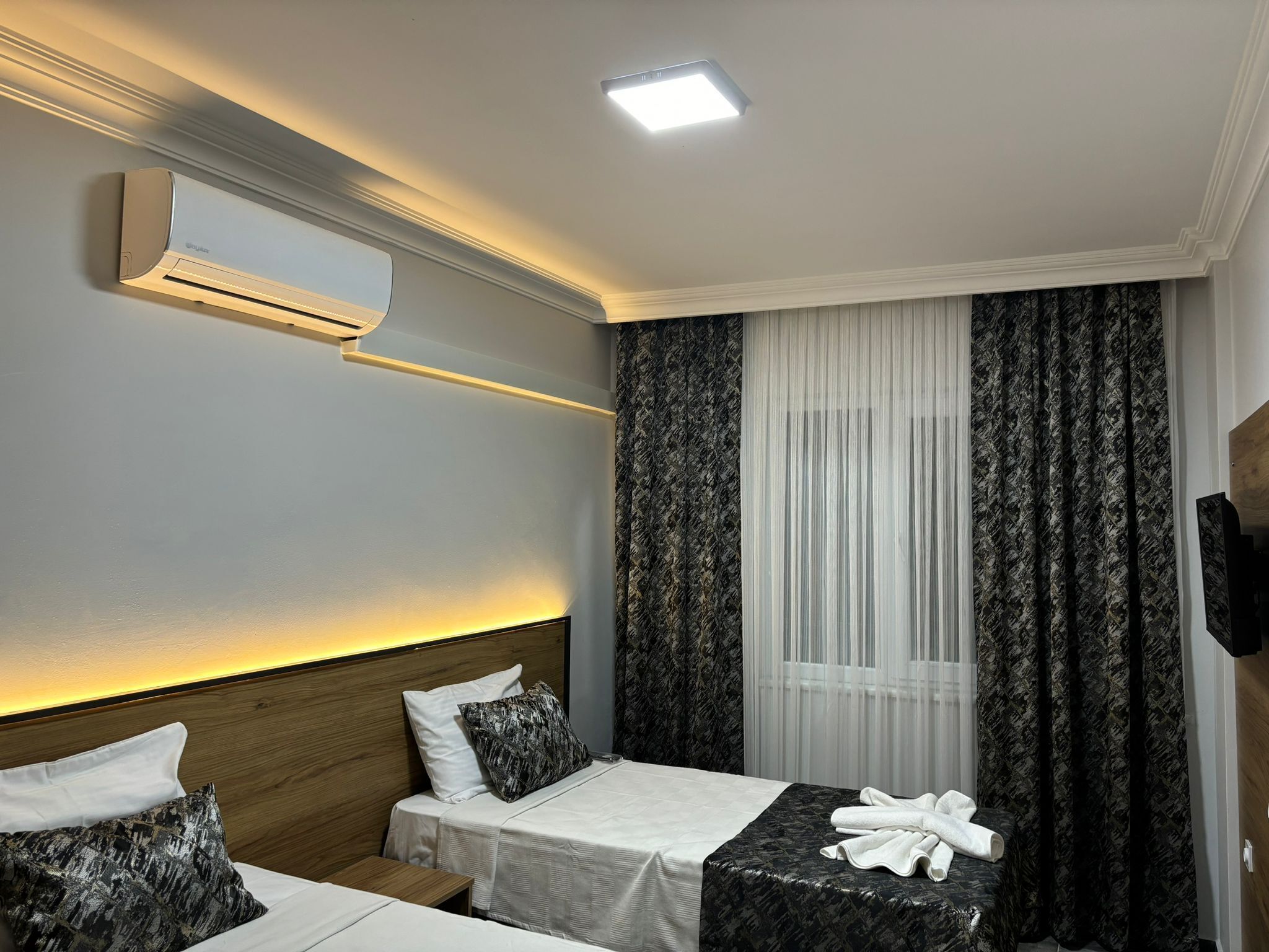 Habitación de hotel con cama, mesita de noche y una pequeña mesa con toallas. Se observa una unidad de aire acondicionado en la pared y una ventana con cortinas.