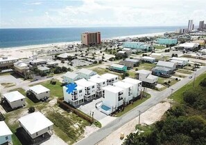 A coastal area with multiple buildings, including a prominent white structure with a blue star symbol, a beach, and a distant high-rise building. - Stunning Luxury Home- WALK to POOL & BEACH! (Gulf Shores)