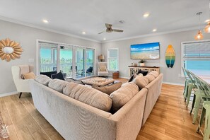 Un salon spacieux avec un grand canapé d’angle beige, un ventilateur de plafond et une vue sur l’extérieur grâce à des portes vitrées.