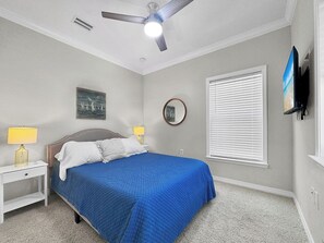 Une chambre à coucher comprenant un lit, deux lampes de chevet, un ventilateur de plafond, une fenêtre avec des stores et un téléviseur fixé au mur.