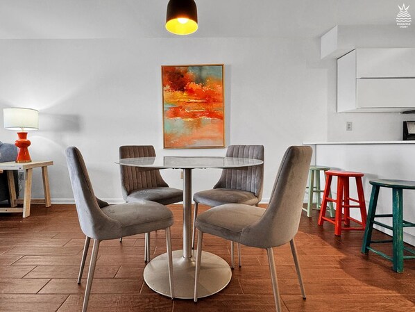 A modern dining area with a round table and grey upholstered chairs, a colorful abstract painting on the wall, and a kitchen area with white cabinets. - Sunset Harbor Condo w/Balcony, Fishing Pier & Pool (Navarre)