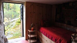 Habitación de cabaña de madera con cama, una mesita con un florero y vista a un área boscosa.