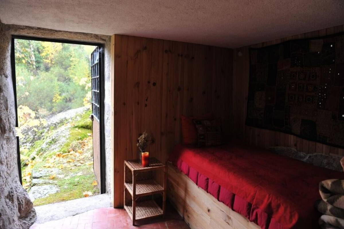 A wooden cabin room with a bed, a small table with a vase, and a view of a forested area outside.