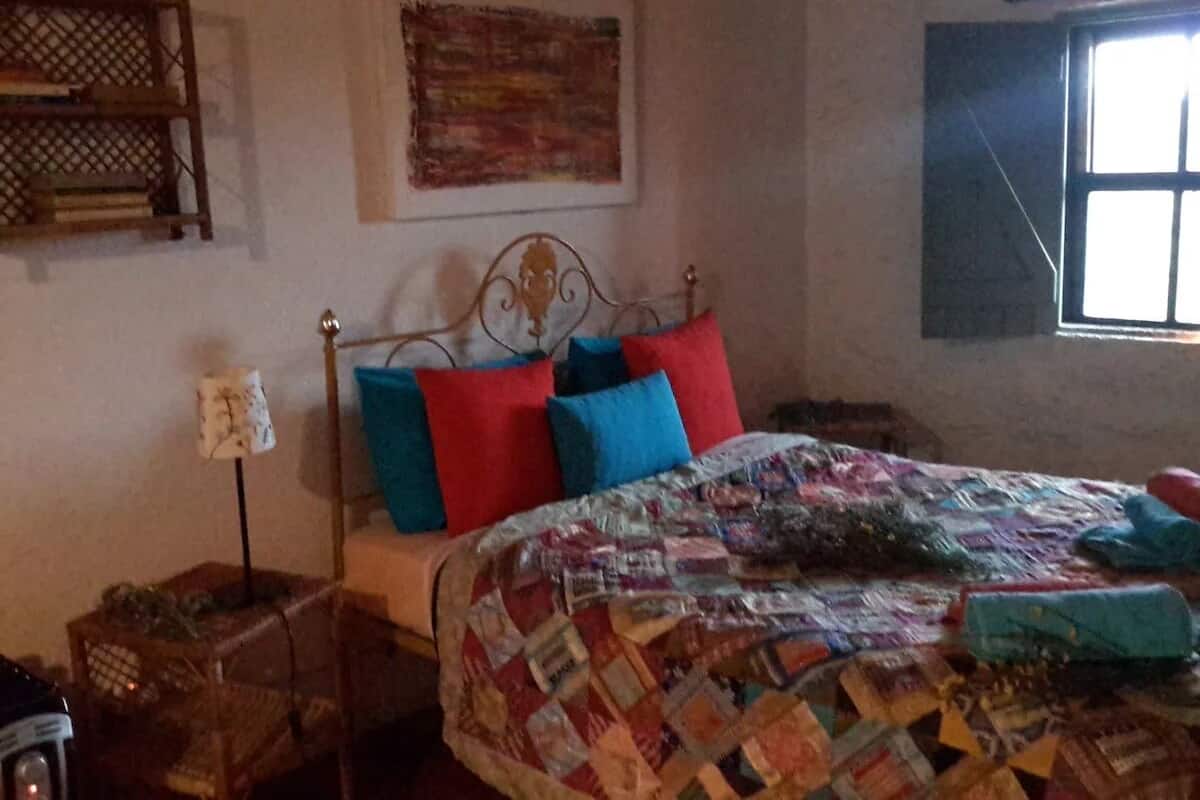 A bedroom with a bed, a nightstand, a lamp, a window, and a wall-mounted shelf.