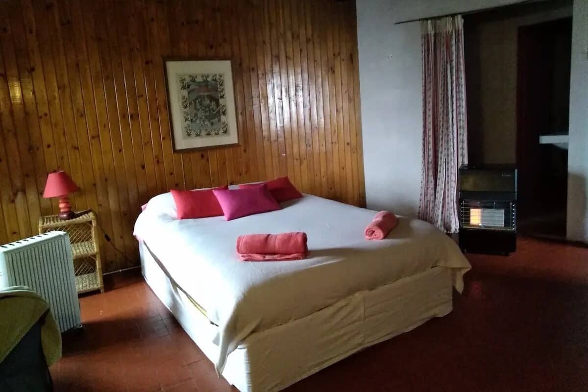 A bedroom with a wooden ceiling, a bed with white and pink bedding, a radiator, and a small table with a lamp.