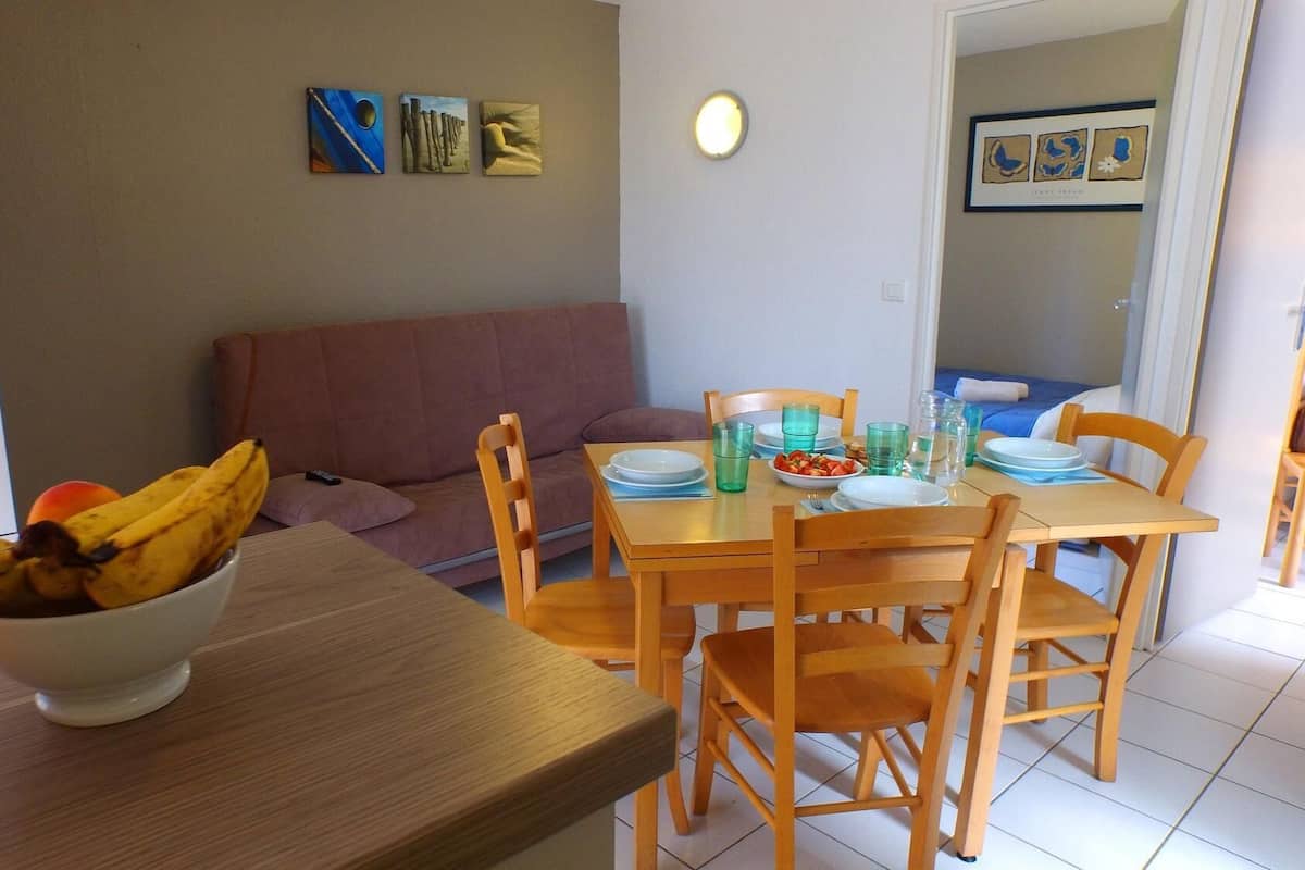 Un comedor con una mesa de madera dispuesta para dos personas, un frutero sobre la mesa y un sofá al fondo.