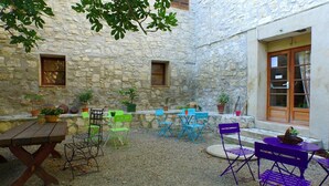 A courtyard with stone walls, a wooden table, and several chairs. There are potted plants and a window with a wooden door. - Vvf Villages Sigean (Sigean)