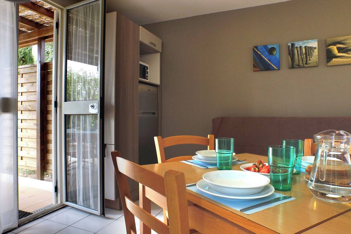 Un comedor con una mesa preparada para dos, un puerta corrediza de vidrio que da a un espacio exterior y un refrigerador al fondo.