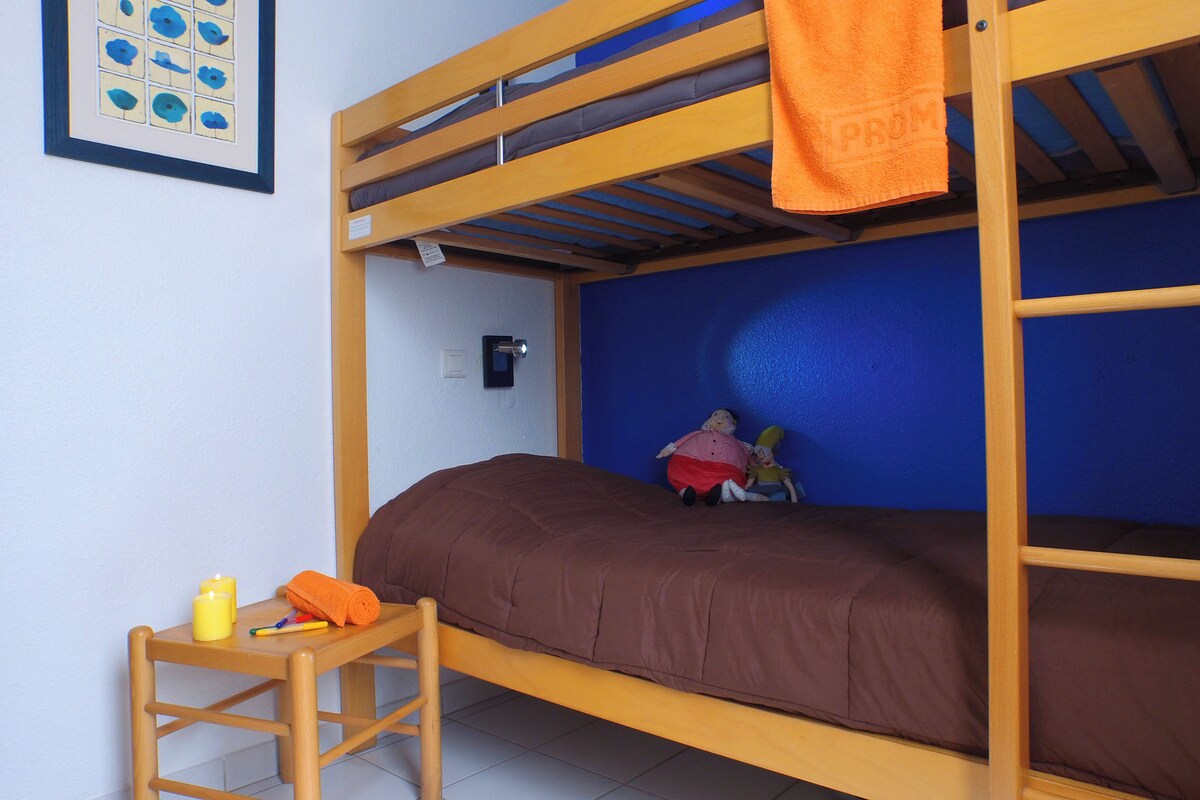 A bunk bed with a brown mattress, a small wooden table with a rolled-up orange towel, and a yellow candle.
