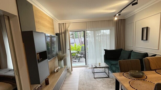 A modern living room with a flat-screen TV, a sofa, a dining table with placemats, and a view of the outdoors through a sliding glass door. - Apartamenty na Wydmach BR (Międzywodzie)
