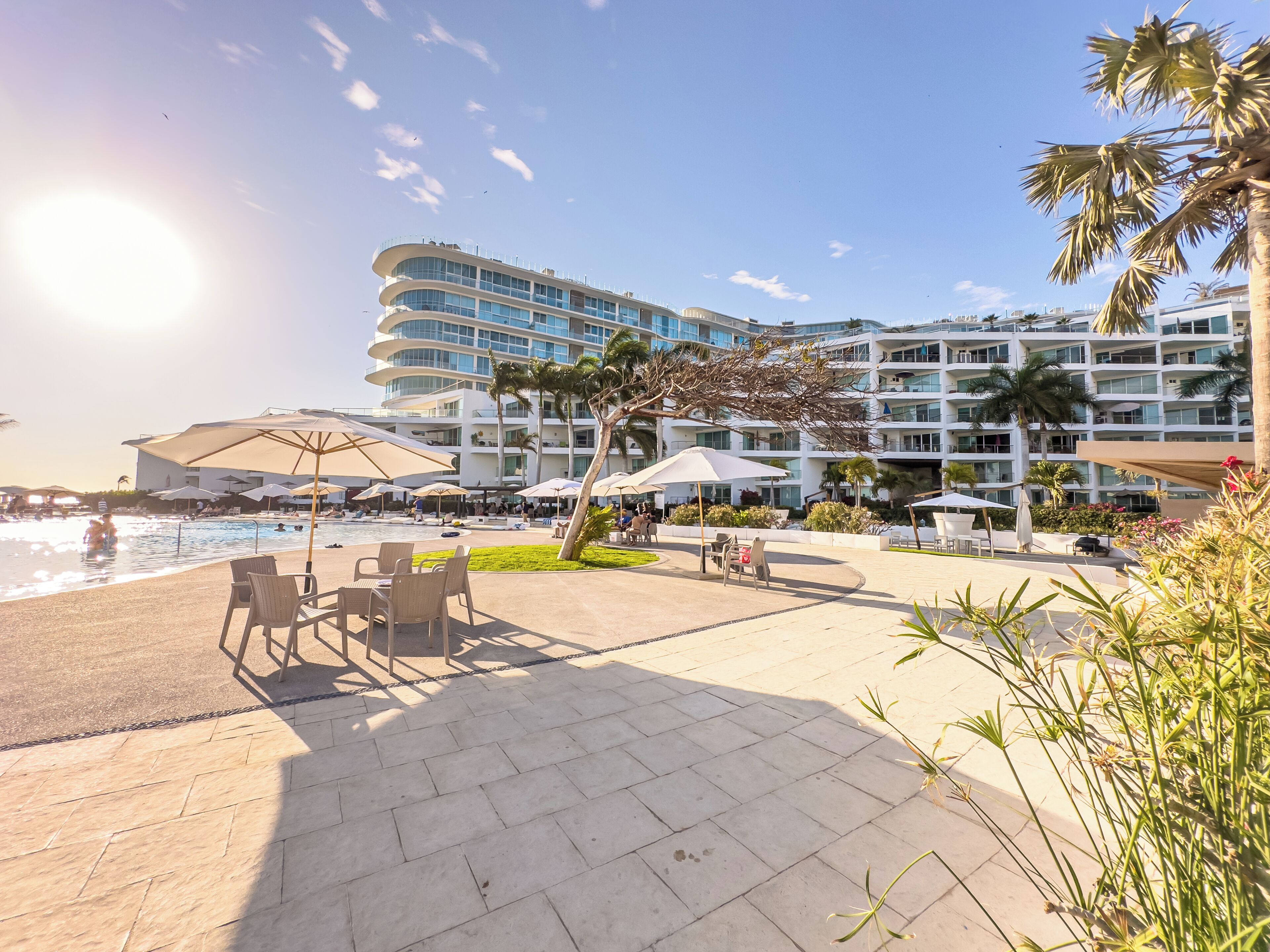 Un área de piscina en un resort, con sillas de descanso y sombrillas, un edificio grande con varios balcones y palmeras.