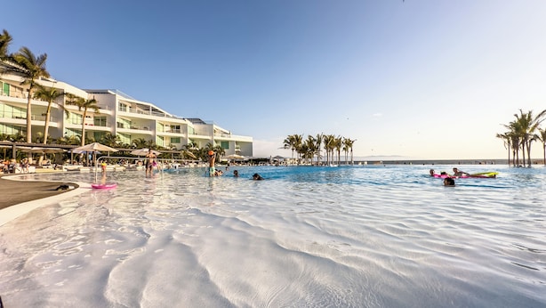 Zona de piscina en un resort, con una alberca grande, palmeras y un edificio con varios balcones.