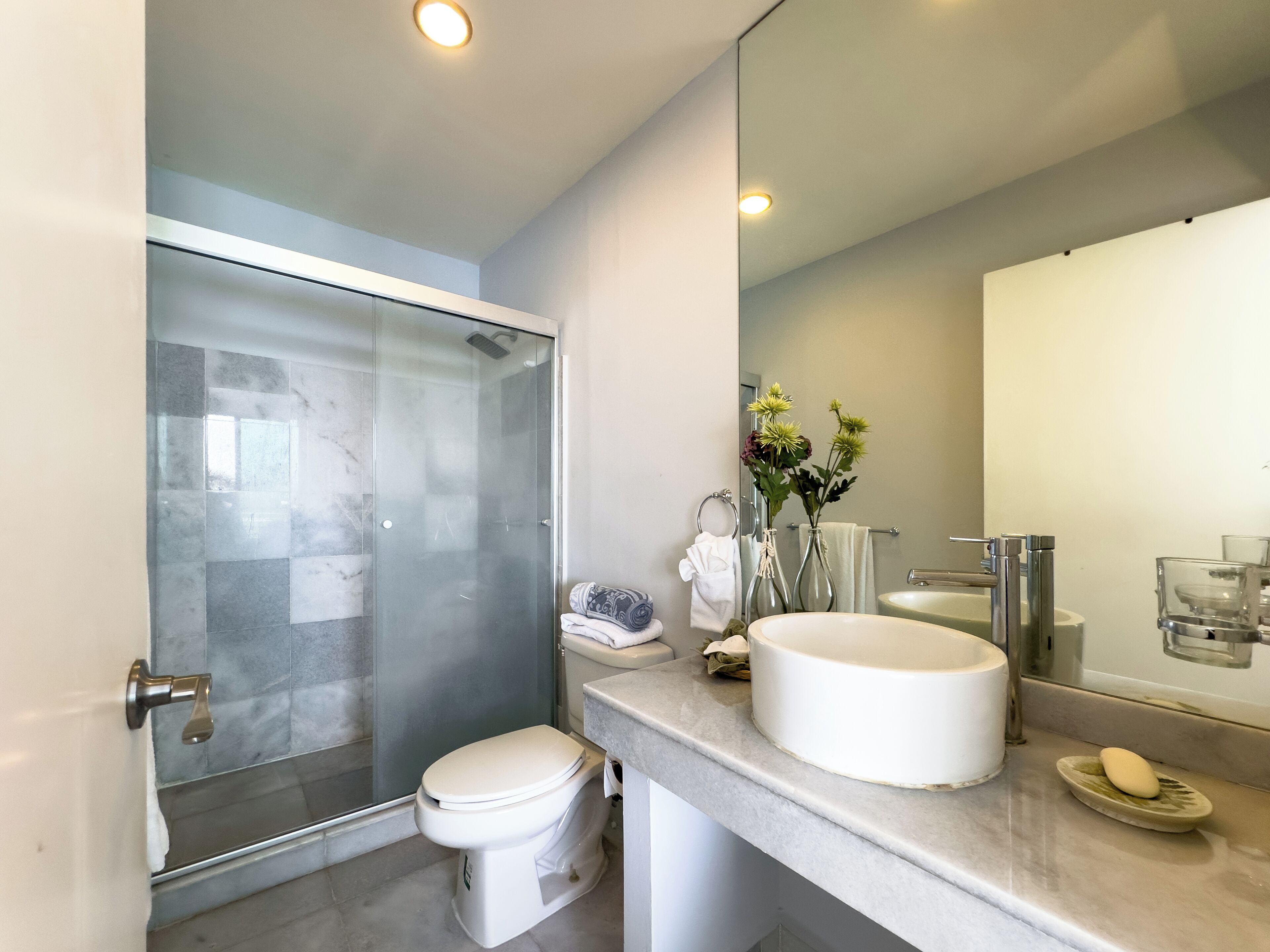 A modern bathroom with a glass shower enclosure, a toilet, a sink with a mirror, and a vase with flowers.