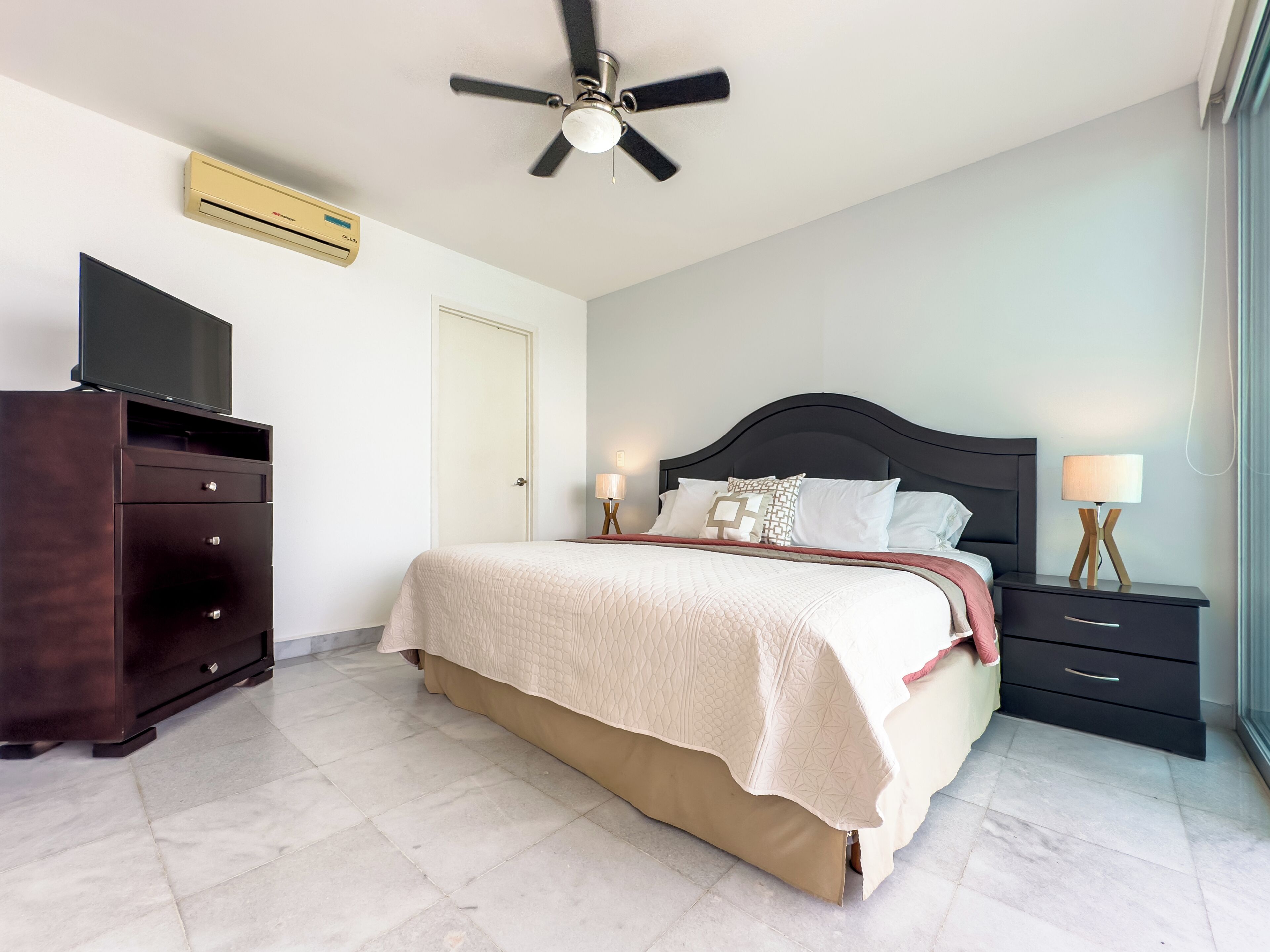Un dormitorio con una cama, ventilador de techo, televisión, cómoda y mesita de noche con lámpara.