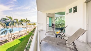 Un balcón con vistas a una piscina y palmeras, una mesa con sillas y un ventanal que da a un espacio interior.