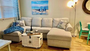 Una sala de estar con un sofá modular, una mesita con una botella y vasos, un cofre decorativo y un cuadro en la pared.
