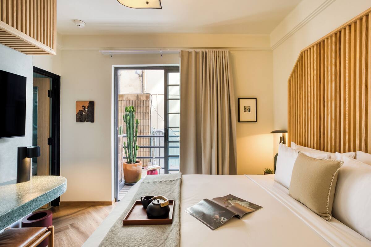 A hotel room with a bed, a TV, a window with curtains, a potted plant, and a tray with a teapot and cups.