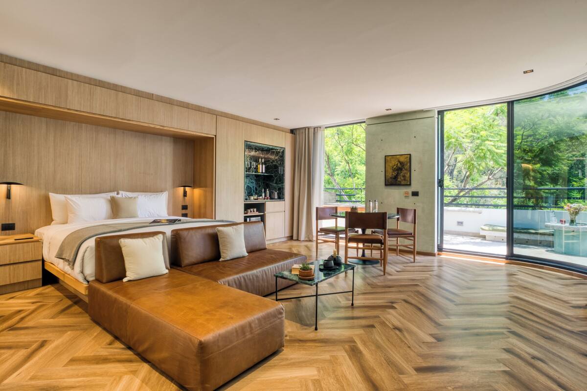 A modern bedroom with a large bed, a sofa, a dining area, and a view of greenery through large windows.
