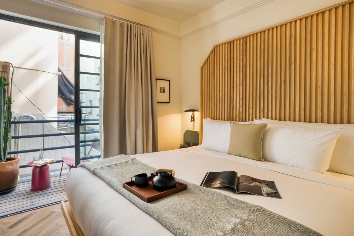 A hotel room with a large bed, a wooden headboard, a window with curtains, a small table with a plant, a tray with tea items, and a magazine on the bed.