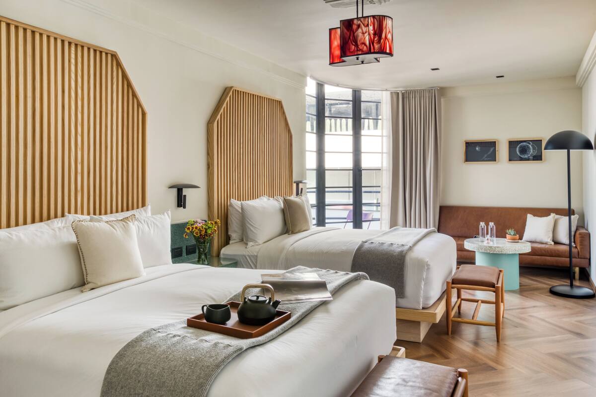 A modern hotel room with two beds, a sitting area, and a large window.