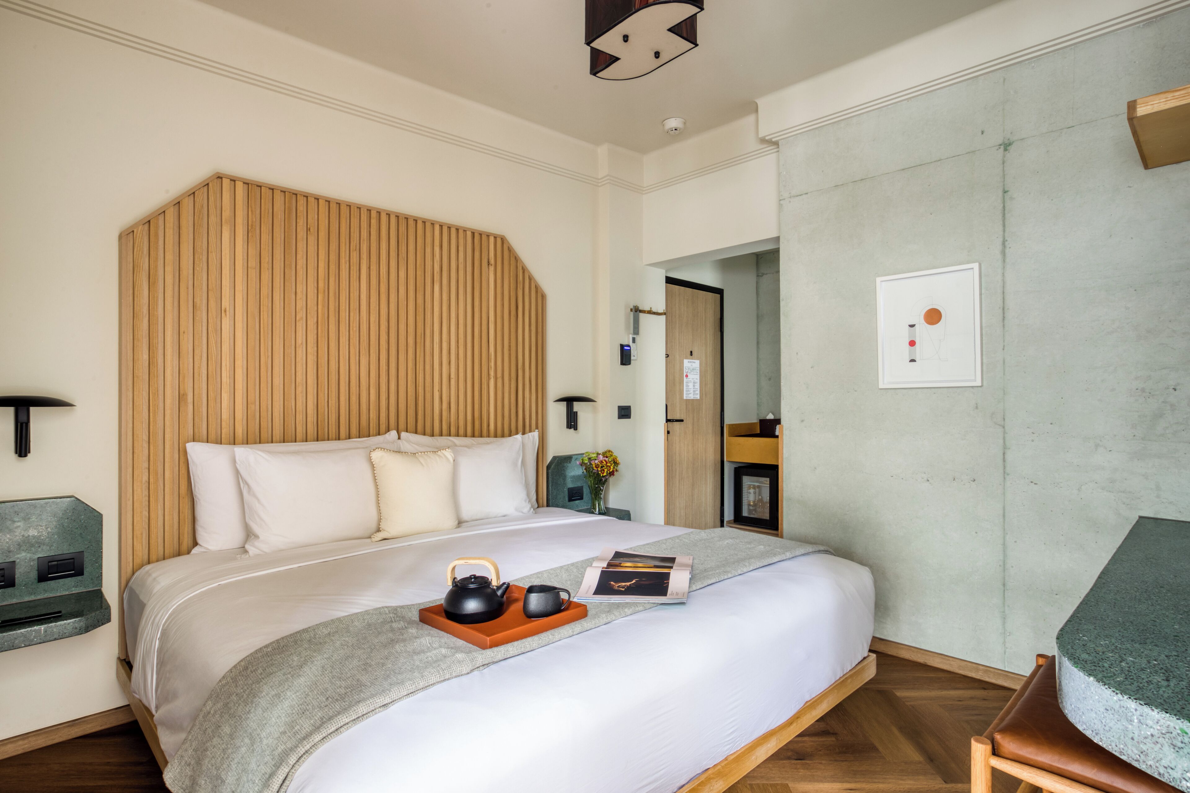 Une chambre d’hôtel moderne avec un grand lit, une tête de lit en bois, une table de chevet avec une lampe et un petit coin salon comprenant une chaise et une table.