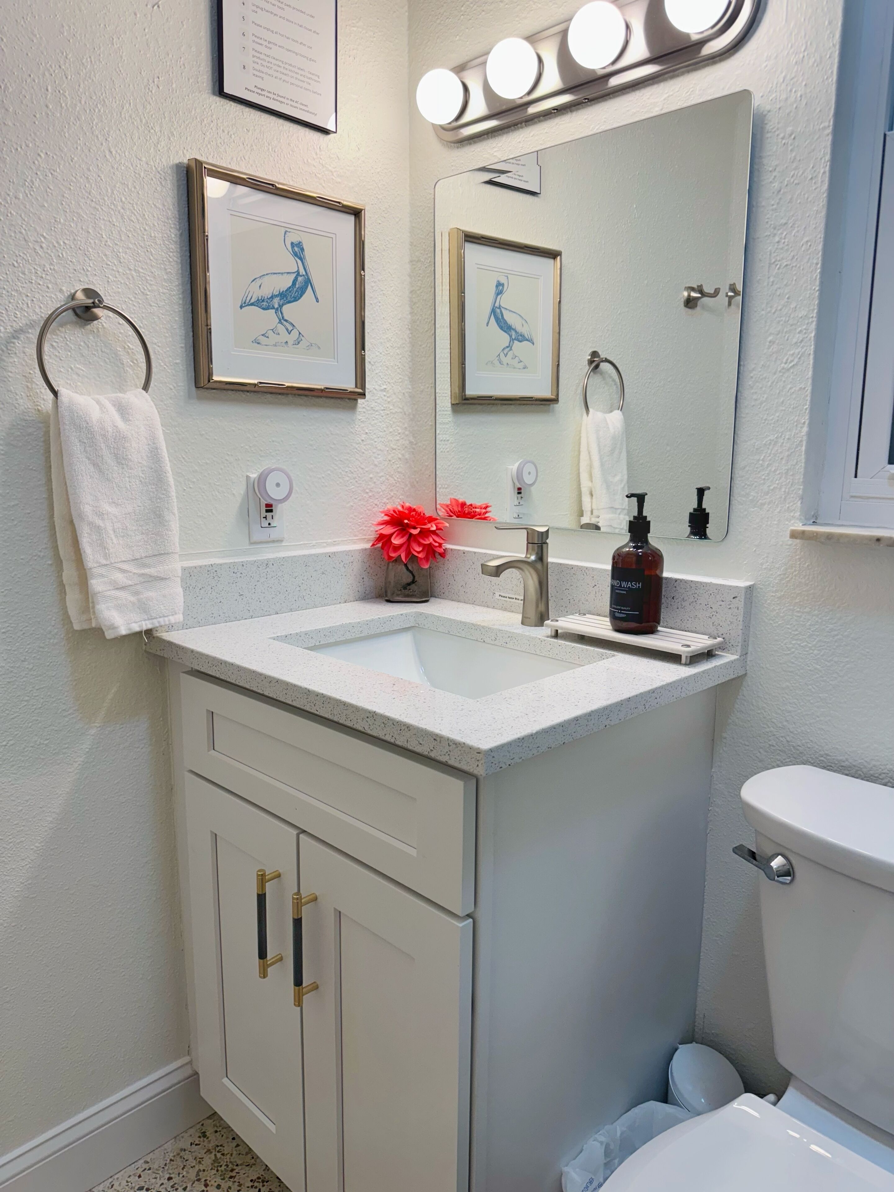 A bathroom with a white sink, a mirror with lights, framed artwork, and a toilet.