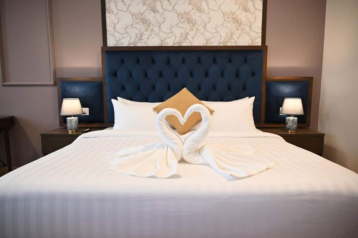 A neatly made bed with a heart-shaped towel arrangement, a tufted headboard, and two bedside lamps on wooden nightstands.