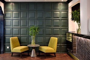 A room with a green paneled wall, two yellow chairs, a round table with a vase of green plants, and a black counter with a mosaic backsplash. - The Leith (George Town)