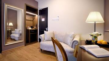 Quarto de hotel com sofá, cadeira, escrivaninha com luminária, espelho e porta para o banheiro.