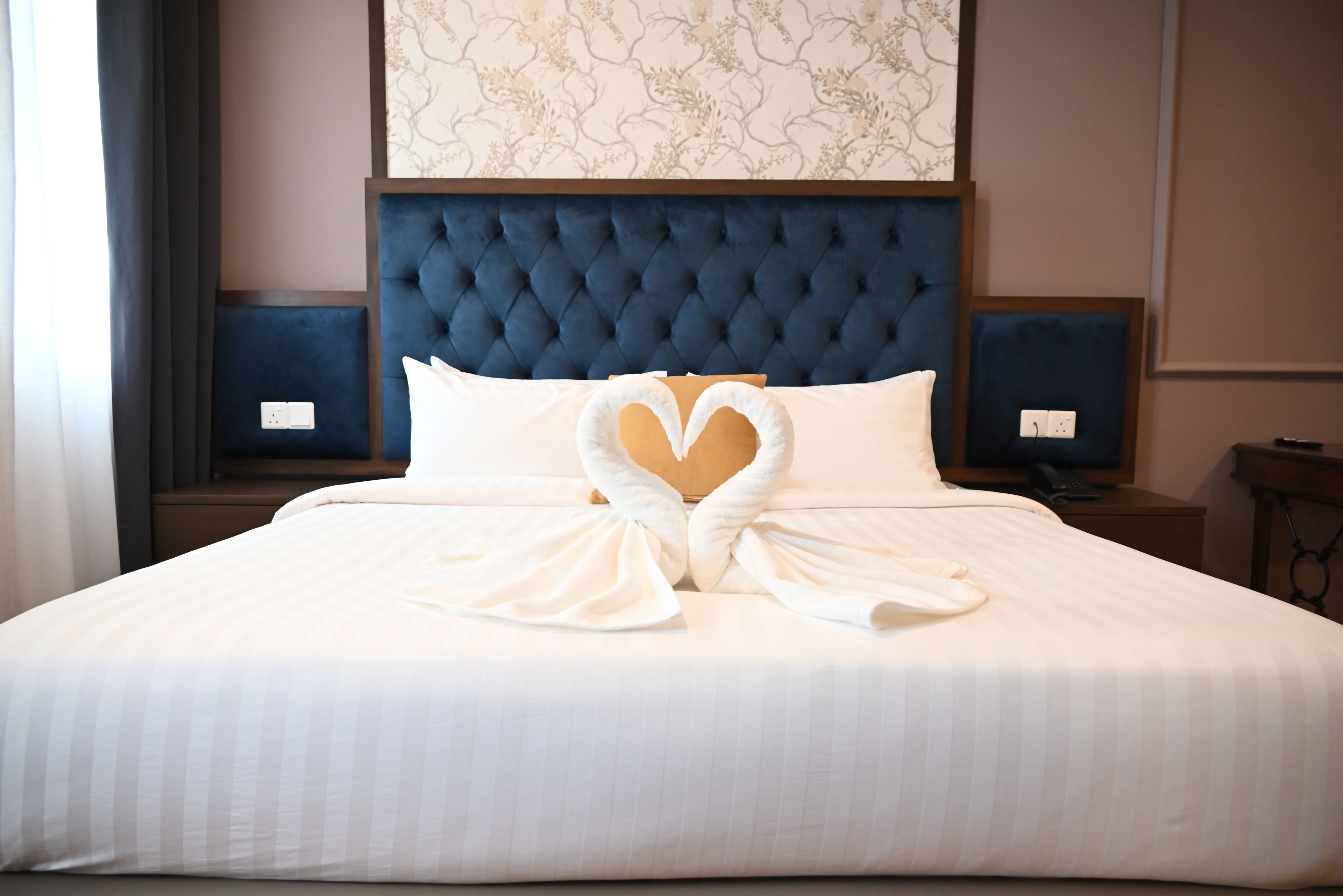 A hotel room with a large bed, two bedside tables, a headboard, and a wall with a floral pattern.