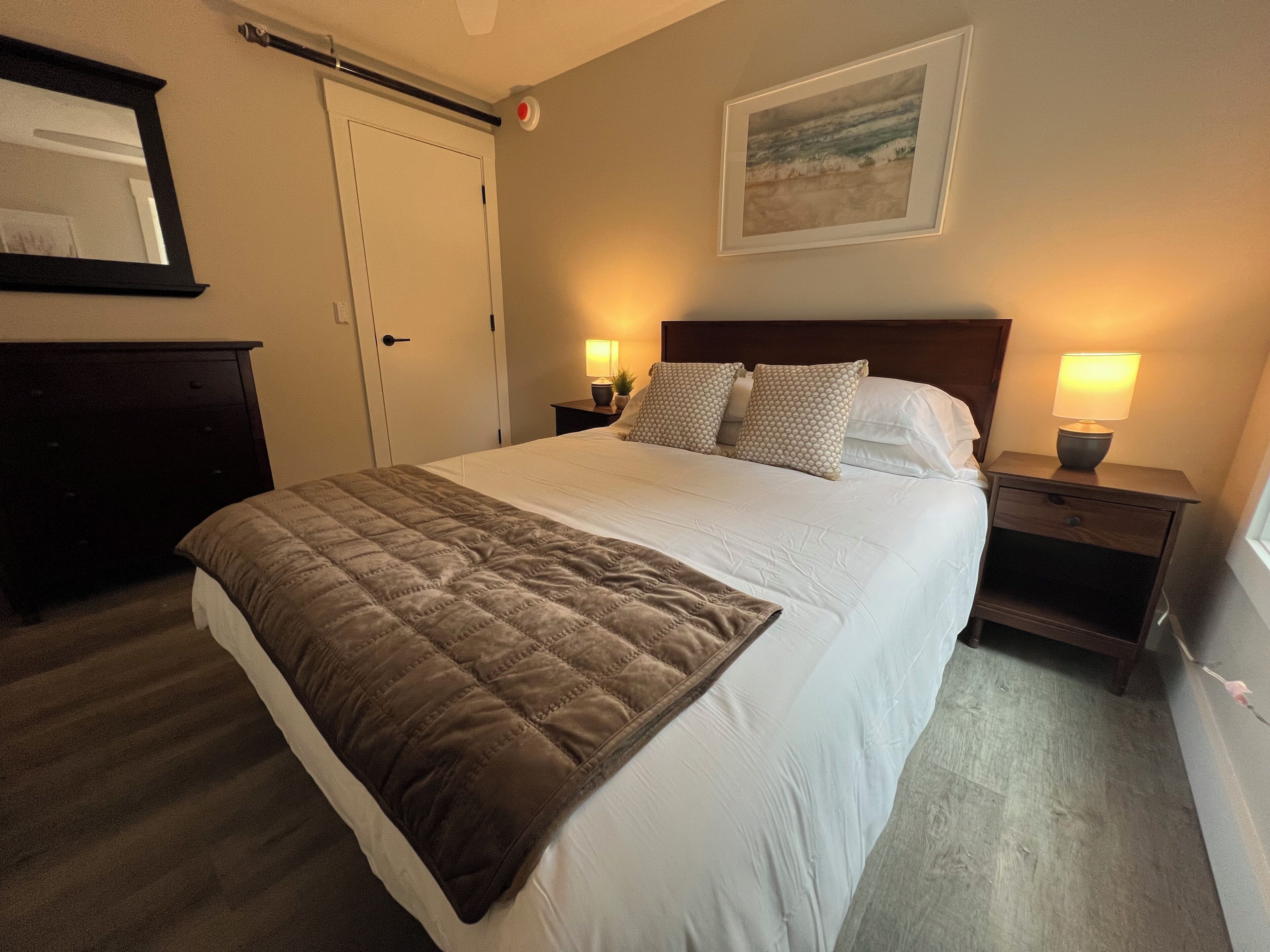 A neatly made bed with a brown quilt, two bedside tables with lamps, a framed picture on the wall, and a door with a red emergency button.