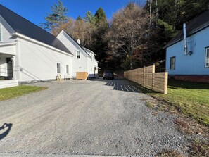 Une allée de gravier mène à un bâtiment blanc, bordé d’une clôture en bois, et on aperçoit un bâtiment bleu au fond.