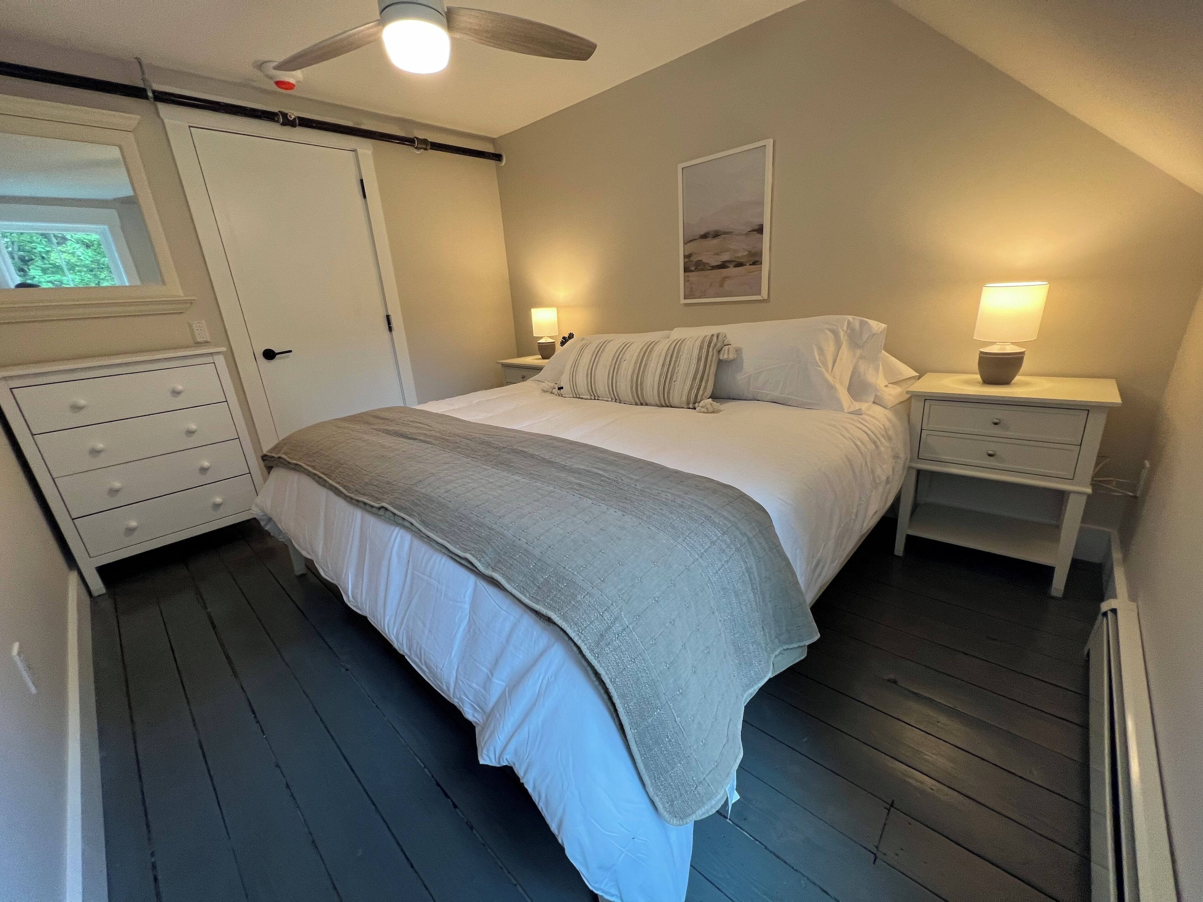 A bedroom with a bed, two bedside tables, a nightstand, a ceiling fan, a window, and a door.