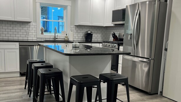A modern kitchen with white cabinets, a stainless steel refrigerator, a gas stove, and a black island with stools. - STAY WITH US, Brand New, 8 BR, 4 Bth, 1/2 mi to Okemo, Hot Tub, on Shuttle, King (Ludlow)
