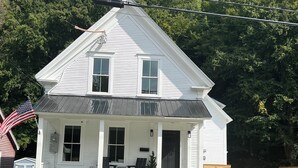 A two-story white house with a front porch, a small patio, and a wooden fence. - STAY WITH US, Brand New, 8 BR, 4 Bth, 1/2 mi to Okemo, Hot Tub, on Shuttle, King (Ludlow)