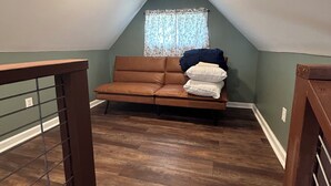 Quarto em estilo loft com um sofá de couro marrom, uma janela com cortinas de renda e piso de madeira.