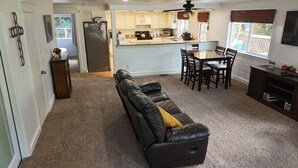 En stue med en sort lædersofa, et spisebord med stole og et køkken med køleskab, ovn og vask.