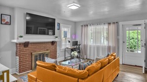 A living room with a brick fireplace, a large flat-screen TV mounted above it, a sofa, a coffee table, and a door leading outside. - Spacious 5BedRooms House Near Millikin Univ – Perfect for Groups & Families! (Decatur)